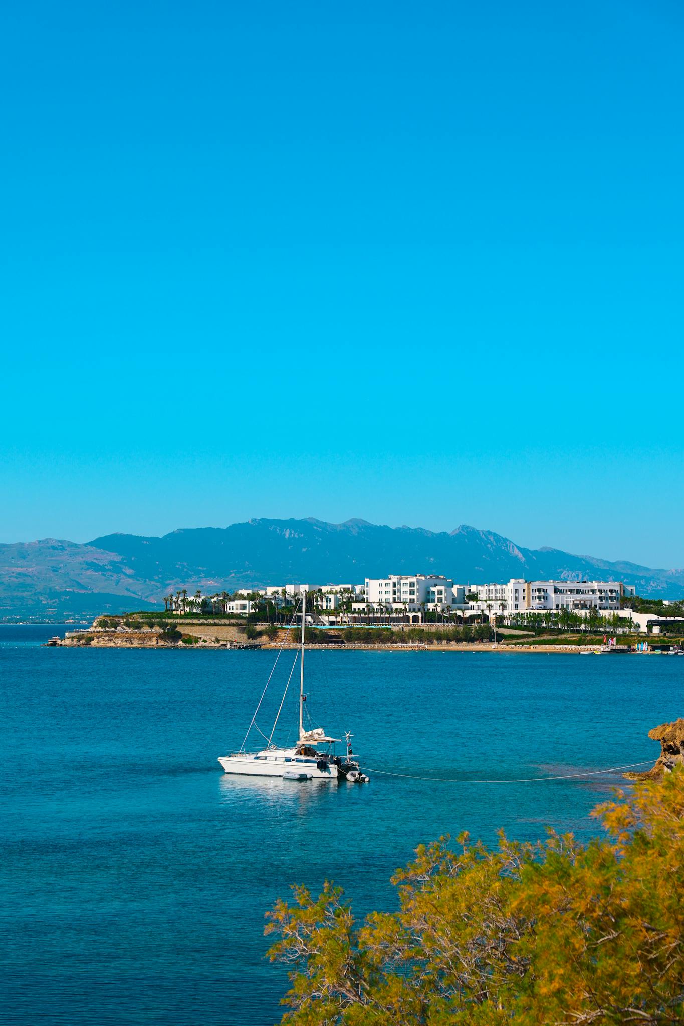 A serene yacht anchored in clear blue waters near a coastal town with a view of distant mountains.